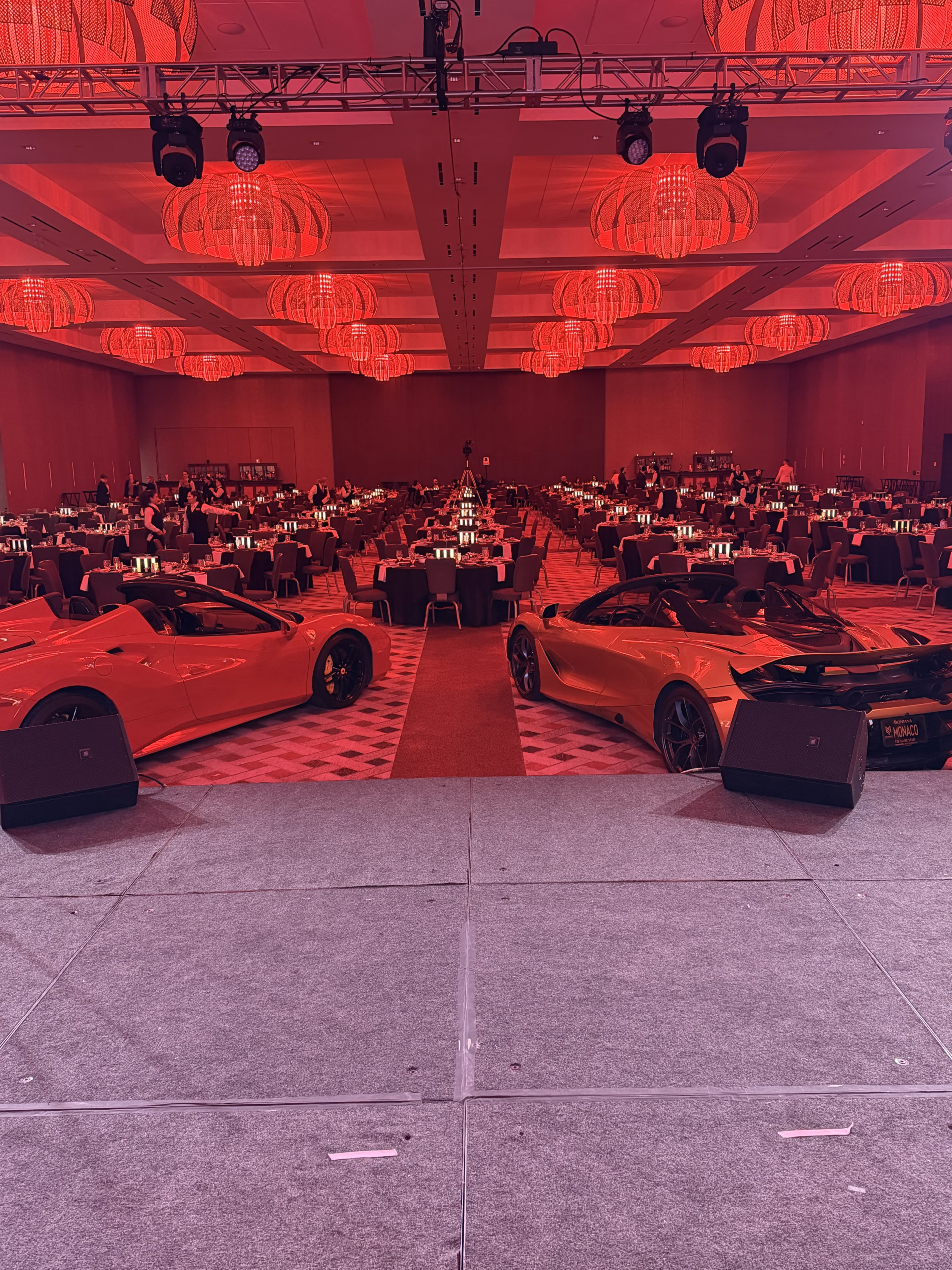 Luxury event ballroom washed in red light, with chandeliers, stage truss, banquet tables, and sports cars