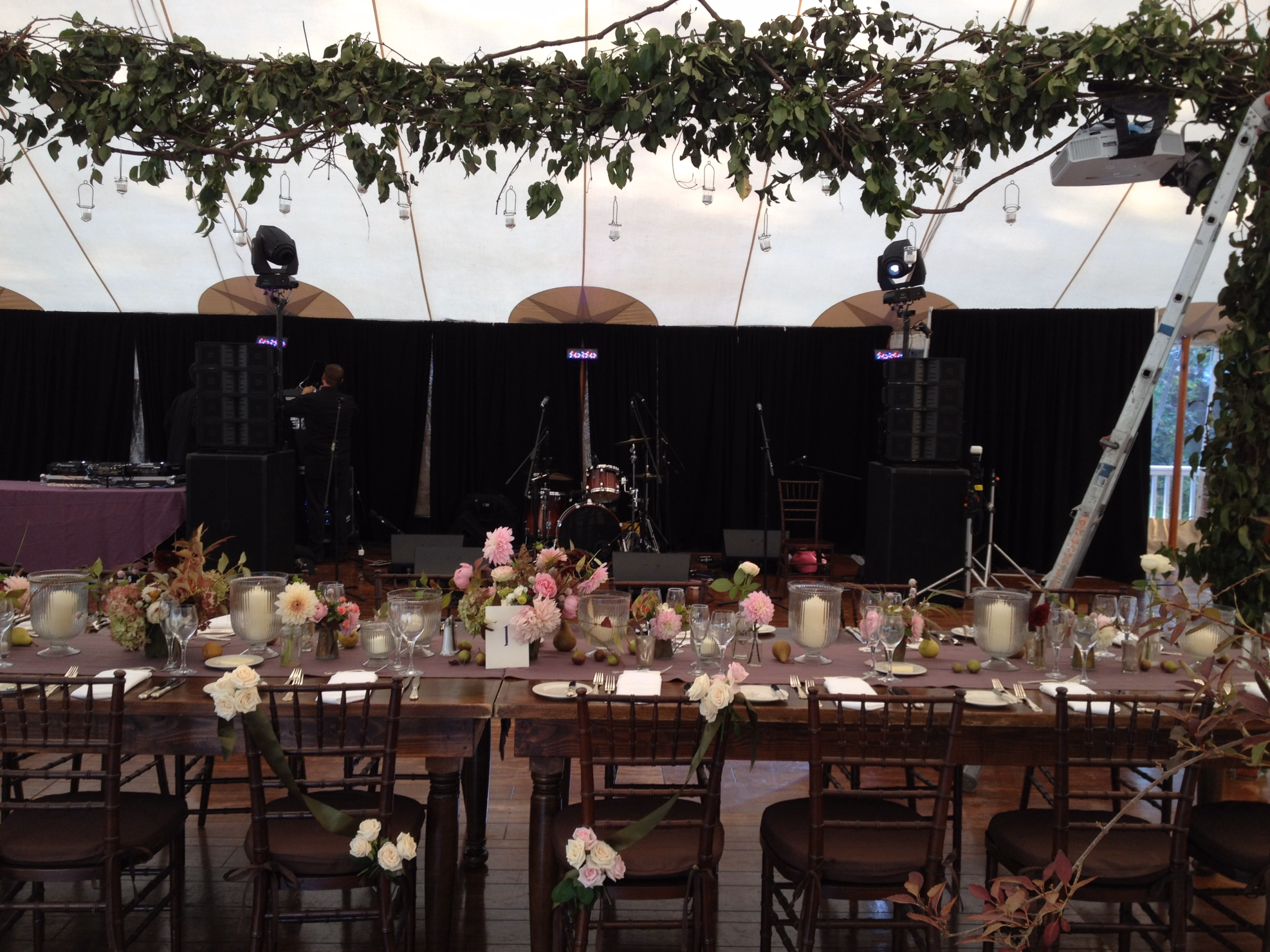 Wedding reception under a tent with leafy overhead decor, floral table settings, audio speakers, and stage lighting