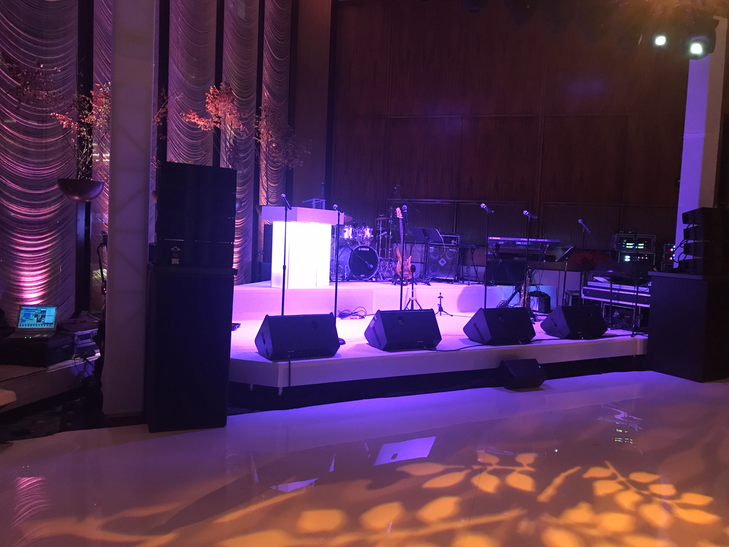 Private event stage with purple lighting, draped walls, audio speakers, microphones, and a dance floor