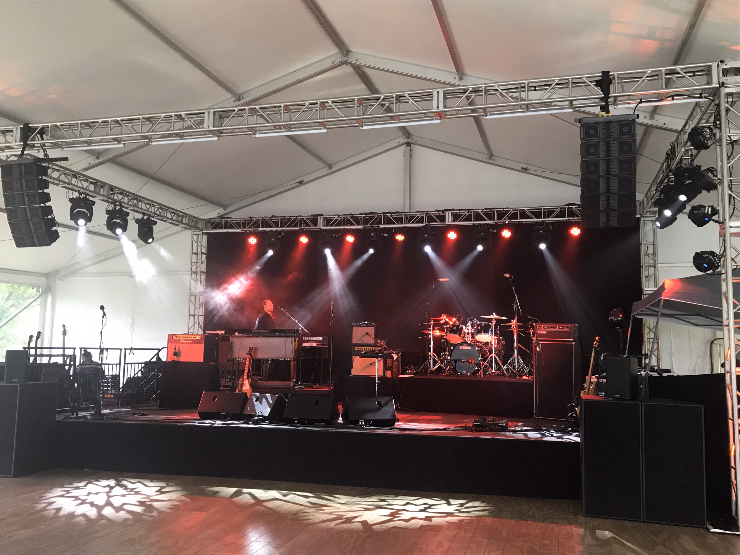 Concert stage under a large event tent with truss, line array speakers, red stage lighting, and band equipment